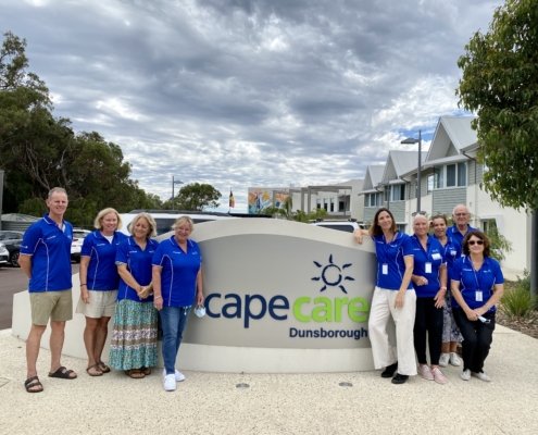Group of 8 people in blue shirts smiling and posing in front of the Capecare Dunsborough entry sign