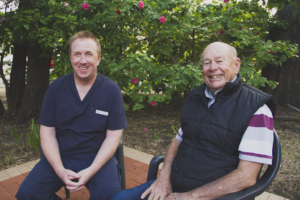Male carer Travis Dean and Male resident David Clarke sitting next to each other outside, smiling at camera. 