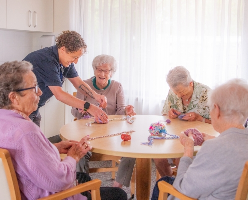 4 older ladies sitting at a table making pom poms with an aged care facility helper