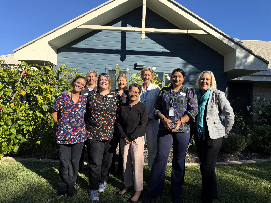 8 professional women of varying ages and nationalities grouped togehter and smiling for the camera outside a blue wall with rose bushes in the background 