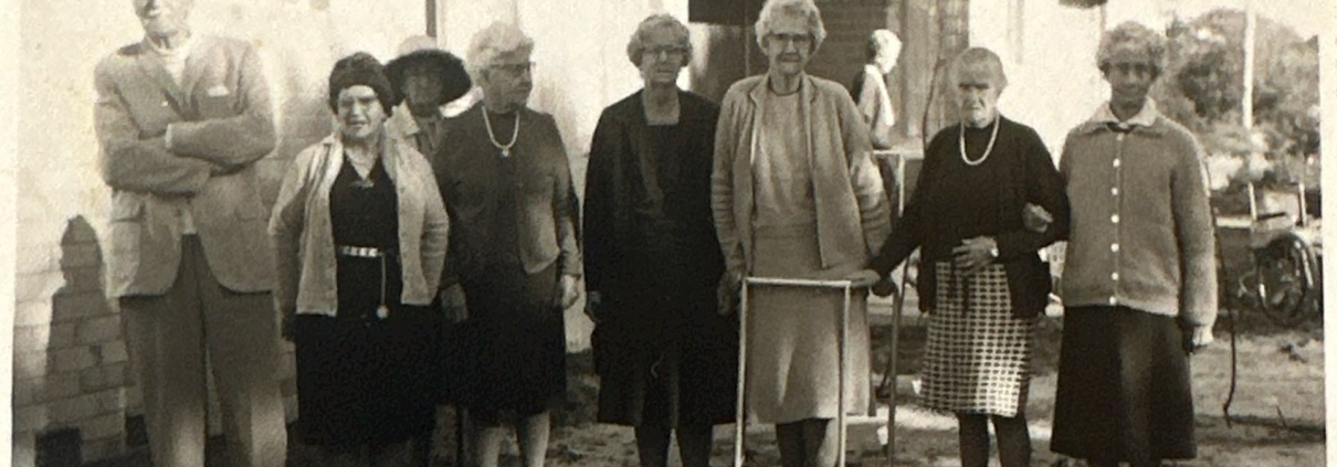 7 elderly women and one man posing for a photograph outside a building. The photograph is sepia and was taken in 1969
