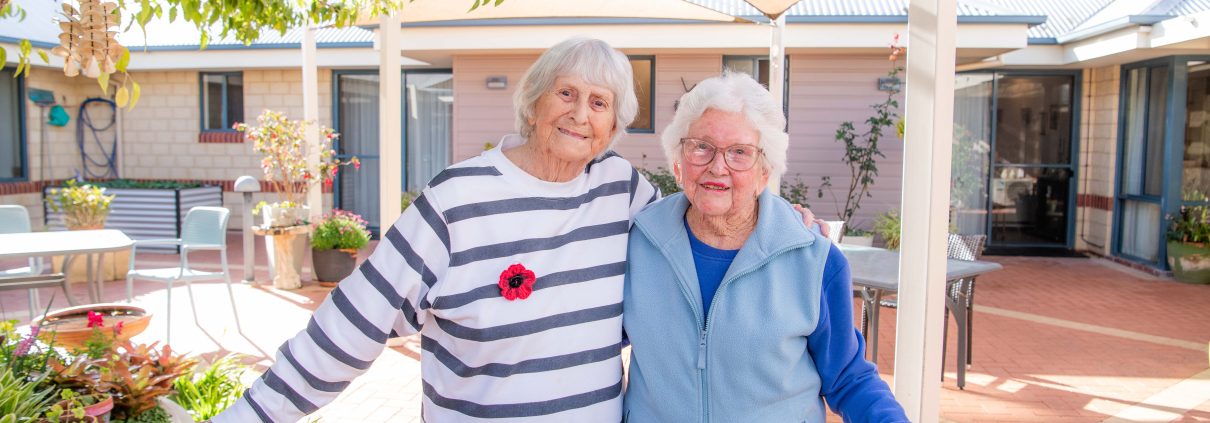 Two older ladies smiling for the camera standing in a courtyard with their arms around eachother's backs