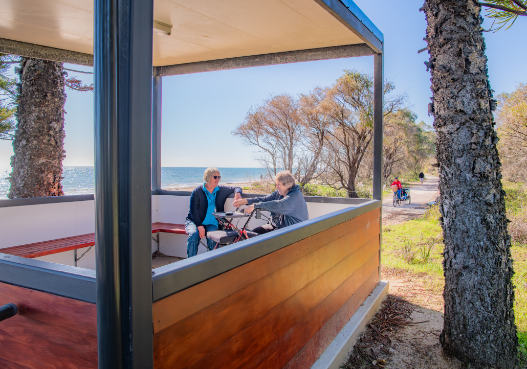 two seated women chatting under a gazebo by a beach pathway on a sunny day